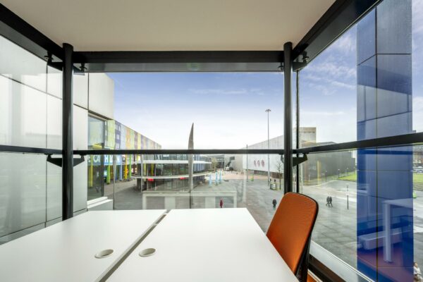Uitzicht vanuit een glazen kantoorruimte op de Johan Cruijff Boulevard met moderne gebouwen en winkelpanden.